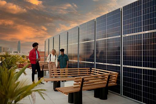 Real estate agent and potential buyers standing on outdoor deck next to solar energy system with dramatic sky and Barcelona cityscape in background at sunset.