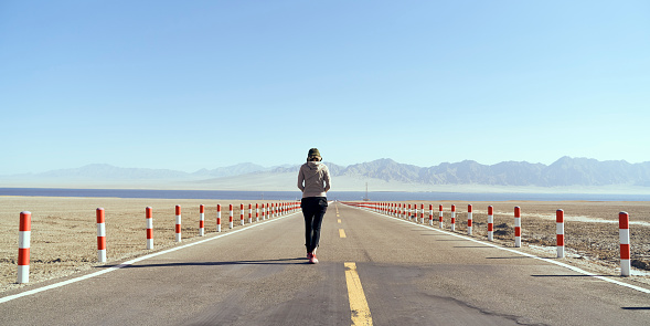 rear view of an asian woman tourist walking on an empty highway towards a lake