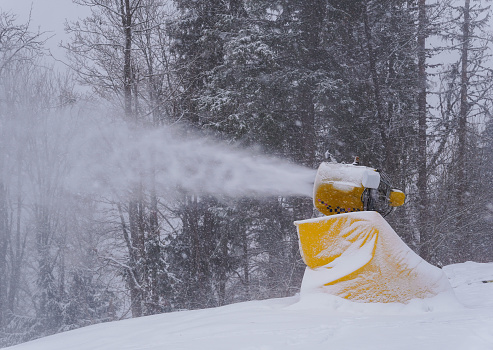Snow cannon during operation. Snow gun spraying artificial ice crystals to ski piste, snowmaking in winter sports resort, trees in background.