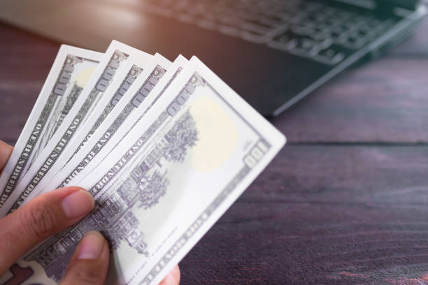 istock Finger holding bank note 100 dollars with keyboard laptop computer blur on wooden table. 1333140007