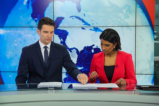 Man and woman sitting at news desk in a tv recording studio. Preparing to go live. Comparing notes.