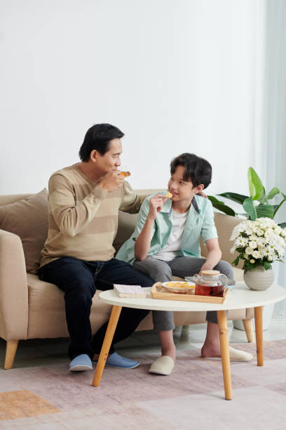 Man Spending Weekend with Son Joyful mature man enjoying spending weekend with son at home, they are drinking tea, eating potato chips and discussing news family eating potato chips stock pictures, royalty-free photos & images