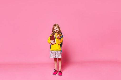 Back to school. Happy blonde girl  child  holds a book,  a red apple in her hand looking at the camera ​on a pink background. Education and intellectual development of children. World book day