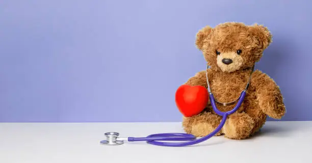 Photo of Teddy bear with a stethoscope and a heart on a purple background. Family doctor or pediatrician concept