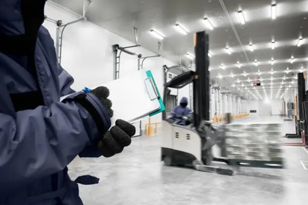 Hand of worker holding clipboard checking goods in freezing room or warehouse with  background of stand-on stacker truck used to lift and move the ready meals goods