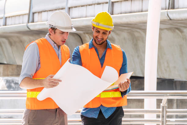 istock Two engineer or architect with safety helmet planning about building with blueprint. Professional technician man pointing finger while his colleagues looking at construction site.Industry work concept 1333160103