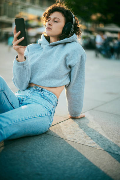 a young woman with curly hair is sitting on the street with headphones on and choosing music on the phone. - typewriter key audio imagens e fotografias de stock
