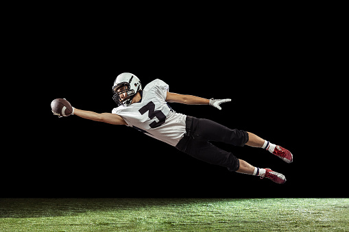 Flying, catching ball. Portrait of American football player training isolated on dark studio background with green grass flooring. Concept of sport, movement, achievements. Copy space for ad