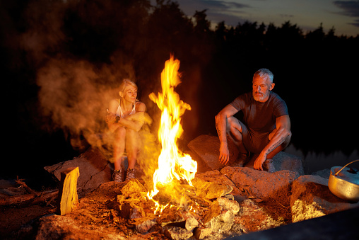 A couple of tourists relaxing near campfire while spending time outdoors in forest. Camping, tourism concept