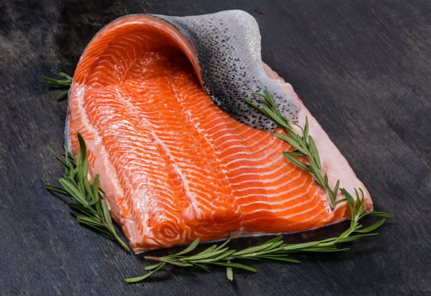Raw trout fillet on skin with rosemary on dark surface Cooled raw rainbow trout fillet in the form of a boneless half of the fish with twigs of fresh rosemary on a dark surface fillet stock pictures, royalty-free photos & images