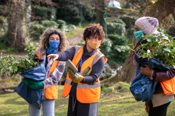 Volunteers Following COVID-19 Guidelines Shot of women wearing protective face masks working together on a community garden outreach project to help improve their local environment. They are wearing hi-vis jackets as they stand together, discussing their plan of action in the North East of England. One woman is using a digital tablet. community garden sign stock pictures, royalty-free photos & images