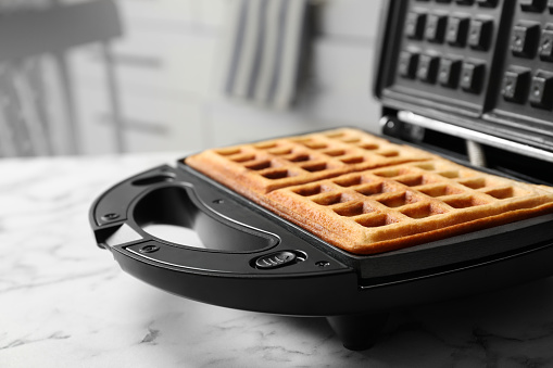 Modern iron with tasty Belgian waffles on white marble table, closeup