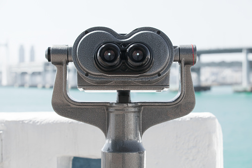 Binocular telescope mounted on a rotating base at the riverside of a modern city. Front view, close up photo with selective focus