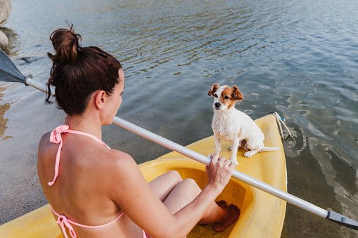 side view of young caucasian woman and jack russell dog sitting on cute jack russell dog sitting on yellow canoe in lake during sunny day. Woman holding oar ready to row.summer time. Pets, adventure and nature