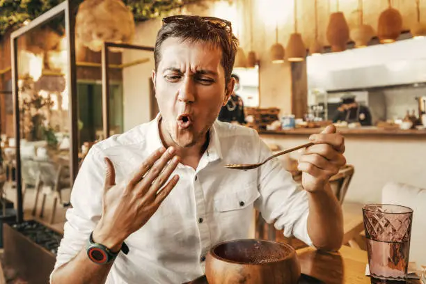 Photo of Funny man tries a spicy and hot dish from the national cuisine. He's all red and is trying to cool his mouth with his hand