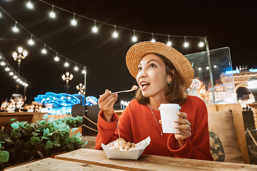 Smiling woman eats a sweet cake with ice cream and drinks coffee in a night cafe with lights