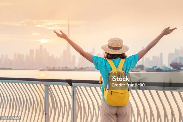 A Happy Traveler Woman With A Hat And A Yellow Backpack Enjoys A Stunning Panoramic View Of The Dubai Creek Canal And The Famous Tallest Skyscraper Burj Khalifa Stock Photo - Download Image Now