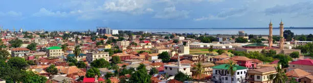 Banjul, The Gambia: city panorama - skyline of the low-rise Gambian capital with the River Gambia estuary and the harbor as background, minarets of the King Fahad Mosque on the right, Atlantic Ocean on the left. The city was built on a shallow sandbar island (St. Mary's Island) that had formed in the estuary over time, it is connected to the mainland via the Denton Bridge. It was mentioned for the first time by Portuguese sailors in the 15th century.