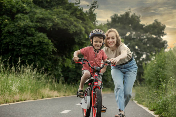 mother teaching son to ride bicycle. happy cute boy in helmet learn to riding a bike on the bike path in summer raining day at sunset time. family weekend - day to sunset imagens e fotografias de stock