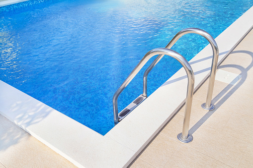 Close up of ladder stainless handrails (stairs) into the blue beautiful swimming pool. Vacation and sport concept.