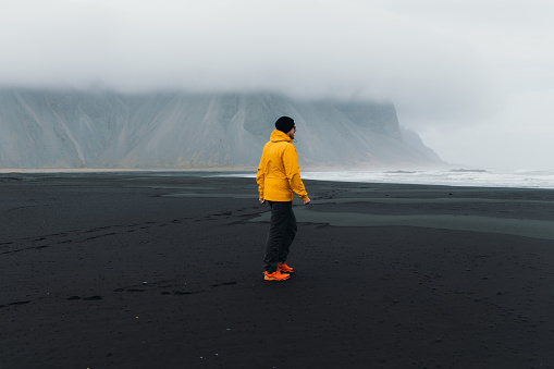 Man tourist in yellow jacket walking at the Stokksnes black beach with view of the beautiful mountain during foggy summer morning in Iceland