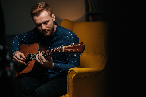 Close-up portrait of handsome guitarist singer male playing acoustic guitar sitting on armchair in dark living room. Creative musician enjoying leisure activity in apartment with modern interior.