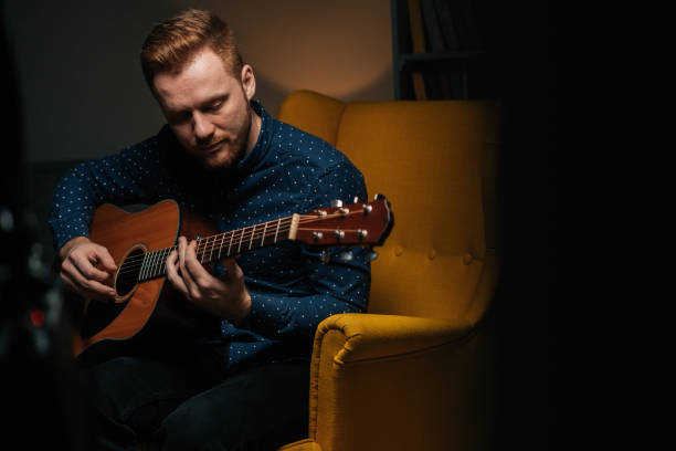 close-up portrait of handsome guitarist singer male playing acoustic guitar sitting on armchair in dark living room. - men artist guitarist guitar imagens e fotografias de stock