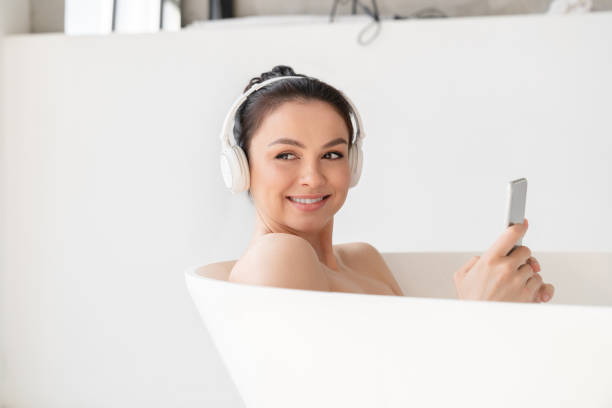 jeune femme satisfaite se relaxant dans une douche chaude et chaude après une dure journée à écouter de la musique dans des écouteurs à l’aide d’un téléphone intelligent. soins de beauté, soins de la peau. concept de procédures de spa - tourist resort audio photos et images de collection