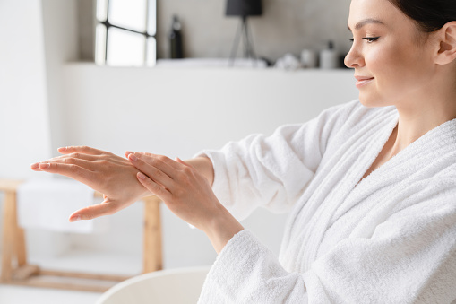 Young caucasian woman in spa bathrobe applying hand cream moisturizer on her hands for softing effect. Recreation, beauty treatment at home