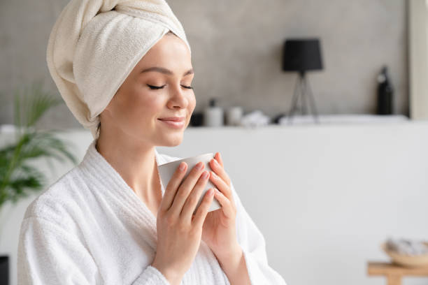 young serene relaxed woman in spa bath towel drinking hot beverage tea coffee after taking shower bath at home. beauty treatment, hydration concept - badjas stockfoto's en -beelden