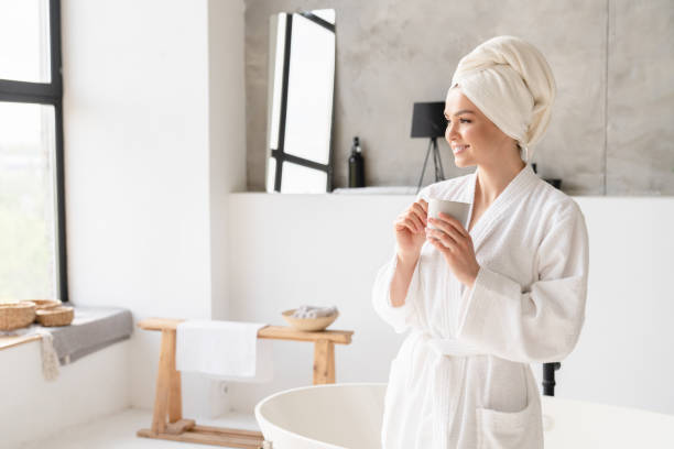 chica joven serena y relajada en toalla de spa de baño y albornoz bebida caliente de agua potable después del baño de ducha en casa. concepto de alivio del estrés. tratamiento de belleza - bathrobe women cup towel fotografías e imágenes de stock