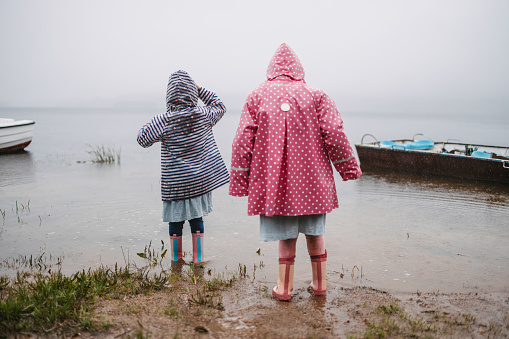 Two girls in colorful raincoats and rain boots stepping into a lake