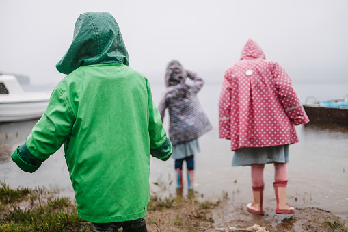 Three children in colorful raincoats and rain boots stepping into a lake