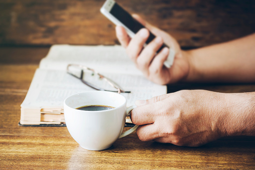 Close up of a man hand holding a cup of coffee and smart phone over holy bible on wooden background