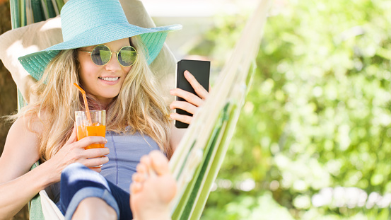 Smiling blonde woman with sunglasses using smartphone, relaxing on the hammock in garden, drinks a juice, free time and summer holiday concept for surf internet or chat with friends using social media