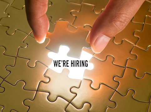 Hand holding a puzzle piece with “We’re hiring” text underneath