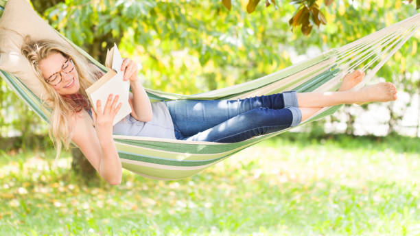 smiling blonde woman with eyeglasses reading a book, relaxing on the hammock in garden, leisure time and summer holiday concept - woman with glasses reading a book imagens e fotografias de stock