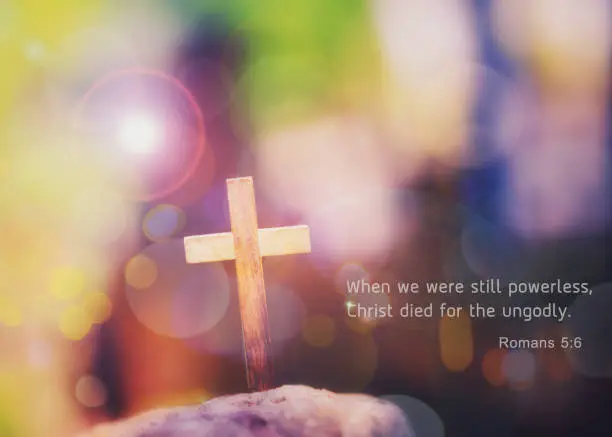 Close up of wooden cross on the rock over blurred colorful  Bokeh  light with word from bible verses, Romans 5:6 , Christian background with copy space.