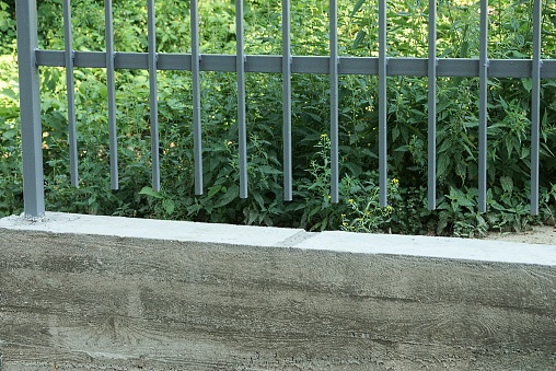 part of a wall of a fence made of a concrete foundation and gray metal rods on the street against a background of green vegetation