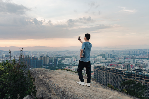 A man using a smart phone to take a photo, the background is on the rock after climbing, facing the city buildings