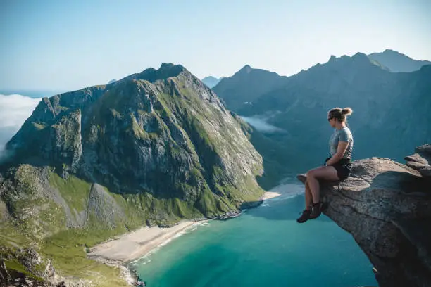 Young woman sitting on cliff in front of blue ocean and mountain in Lofoten, Norway