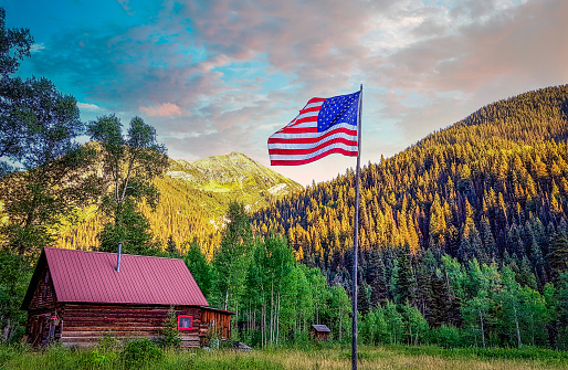 A single flagpole with the American flag in a calm valley with a cabin and a mountain range in the background during sunset.
