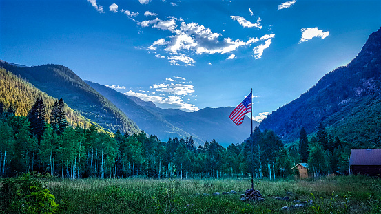 A single flagpole with the American flag in a calm valley with a cabin and a mountain range in the background during sunset.