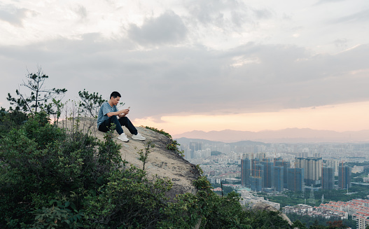 After climbing successfully, an Asian man sits on a rock and shares his joy with his friends. The background is a group of urban buildings.