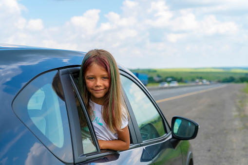 Cute kid girl smiling and having fun to travel. Child look out from car window in countryside. Family travel concept by car on holidays. Family road trip