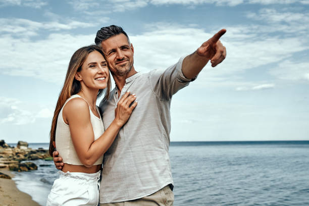 feliz pareja de hombres y mujeres caminando por la playa y hombre apuntando a algo. - couple walking old middle fotografías e imágenes de stock