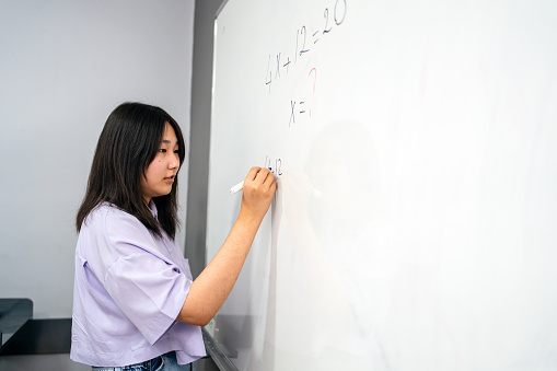 Portrait of Japanese Ethnicity girl  writing solution of sums on white board at school.  Japanese Ethnicity  schoolgirl solving addition sum on white board during Covid-19 pandemic. School child thinking while doing mathematics problem .