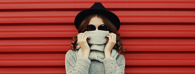 Portrait of young woman covering her face with scarf wearing a black round hat, gray sweater on a red background