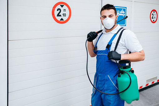 Manual worker in protective work wear spraying virus and bacteria disinfection in underground garage of residential building with sprayer.
