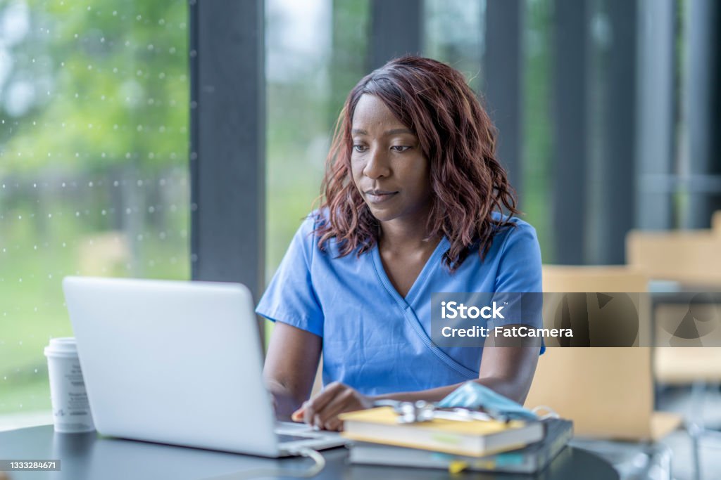 Medical student studying A woman works diligently at her laptop. She is wearing medical scrubs and working in an institutional building. Nurse Stock Photo